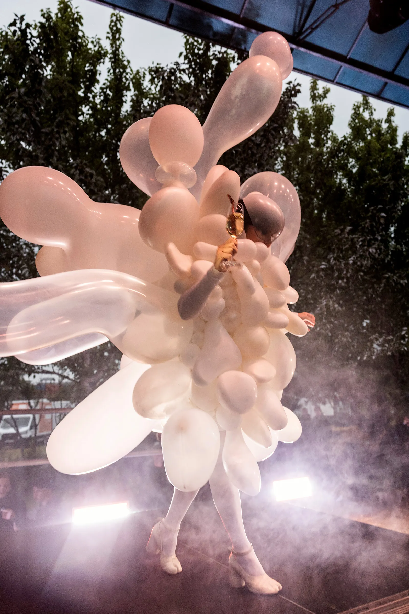 Björk in a balloon costume holding a glass on an outdoor stage.